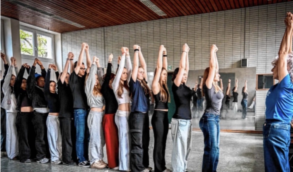 Probe im Tanzsaal: Schülerinnen und Schüler stehen in Reihen mit erhobenen Armen, Aalto-Choreografin Adelina Nigra gibt vorne die Bewegung vor