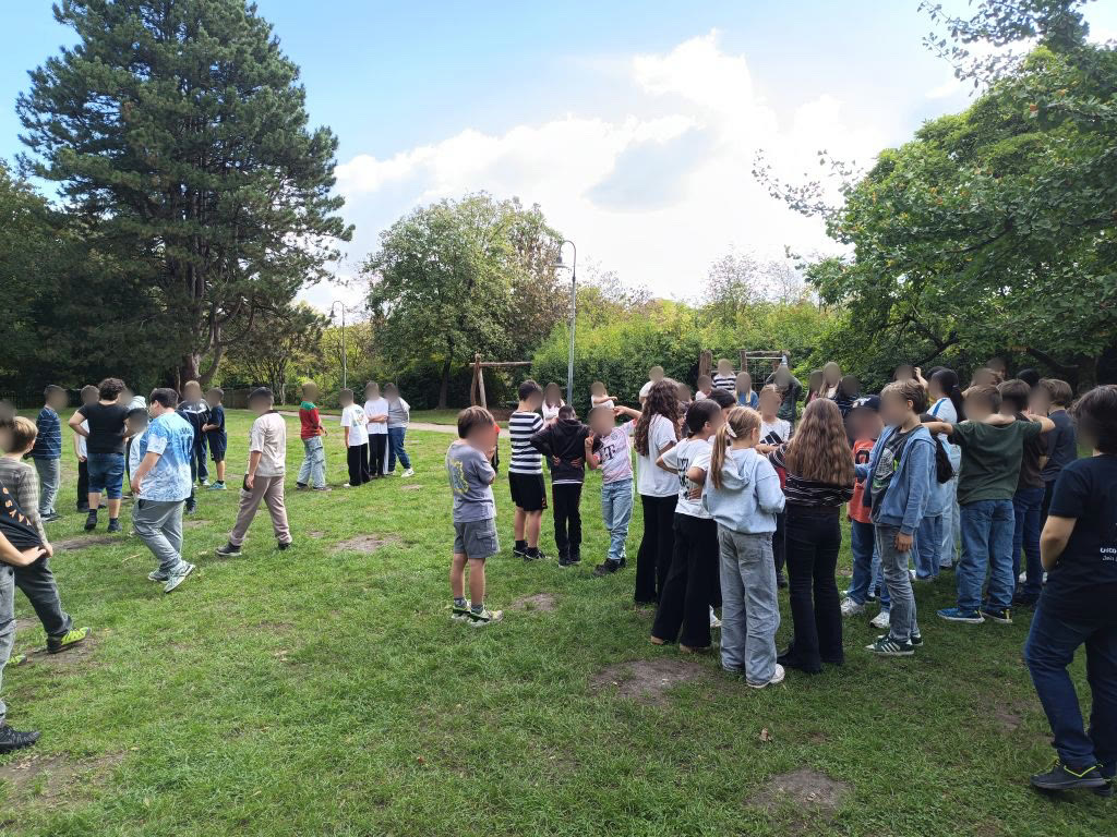 Schülerinnen und Schüler der Klassen 5a und 5b stehen in Gruppen auf einer Wiese im Park, einige legen sich gegenseitig die Arme um die Schultern. Die Gesichter sind weichgezeichnet.