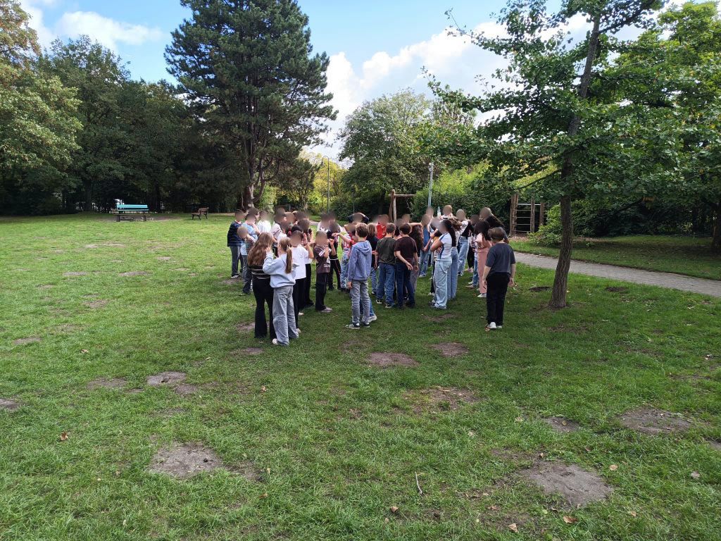 Die beiden fünften Klassen stehen als große Gruppe dicht beieinander auf einer Wiese, im Hintergrund Bäume und ein Spielgerät aus Holz. Die Gesichter sind weichgezeichnet.